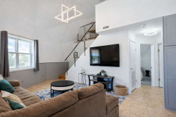 Modern loft-style living room with vaulted ceilings, plush sectional sleek staircase. Natural light fills the space, highlighting neutral tones, geometric lighting & cozy textures—perfect for relaxing, entertaining in your stylish short-term retreat.