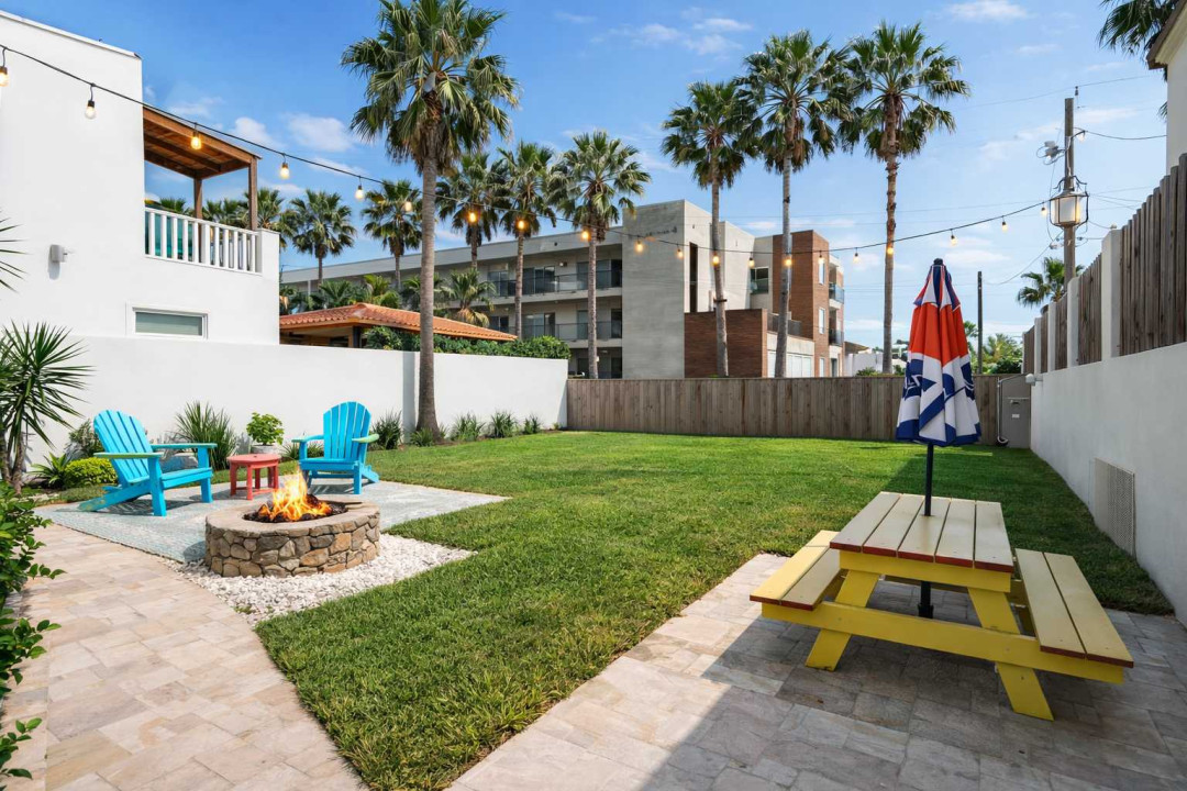 Sunlit coastal retreat with lush lawn, vibrant picnic setup & cozy fire pit under palm-lined skies—perfect for relaxed outdoor dining or evening gatherings. Modern fencing, string lights & tropical accents create a serene resort-style backyard escape