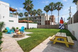 Sunlit coastal retreat with lush lawn, vibrant picnic setup & cozy fire pit under palm-lined skies—perfect for relaxed outdoor dining or evening gatherings. Modern fencing, string lights & tropical accents create a serene resort-style backyard escape