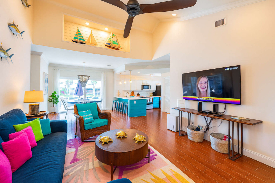 Vibrant living space with plush sectional, colorful pillows, coastal decor, and large TV. Enjoy a spacious open layout that flows to the dining and kitchen area, perfect for relaxing or entertaining.