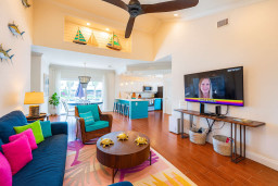 Vibrant living space with plush sectional, colorful pillows, coastal decor, and large TV. Enjoy a spacious open layout that flows to the dining and kitchen area, perfect for relaxing or entertaining.