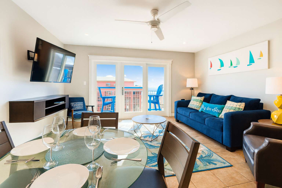 Bright and inviting condo living space with a smart TV, dining table for four, cozy seating, and a private balcony with ocean peeks. Perfect for relaxing after a beach day, enjoying family meals, or unwinding with board games and movies.