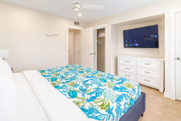 Bright and spacious bedroom with a comfy bed, vibrant coastal bedding, flat-screen TV, dresser, and closet. Ceiling fan and natural light ensure a comfortable stay, blending relaxation with convenience.