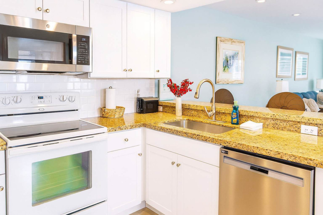 Bright Coastal Kitchen Granite Counters & Stainless Appliances.
Spacious white cabinetry, gold-flecked granite, microwave/oven/dishwasher shine under soft light. Red flowers, beach art & bar stools invite family meals—cook up vacation magic in style.