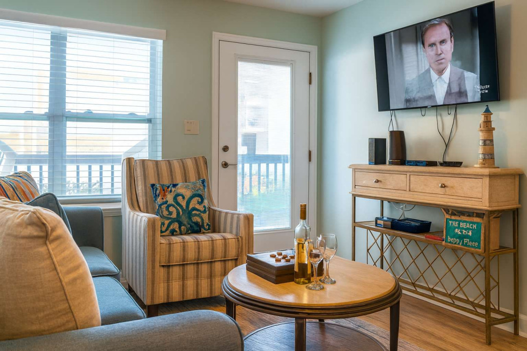 Cozy Beachy Living Room
Smart TV, Bar & Balcony Views. Soft teal walls, plush sofa & patterned chair face wall-mounted TV over driftwood console w/ lighthouse decor & drinks. Slider to balcony—chill, sip & stream in nautical comfort!