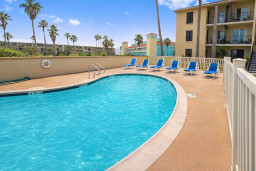 Relax by the sparkling pool at this tropical coastal retreat, featuring blue loungers, palm trees, and vibrant pastel architecture—perfect for sun-soaked getaways and family fun near the beach.
