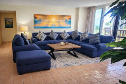 Spacious coastal retreat with vibrant sunset art, plush blue sectional, and balcony views of palm trees. Perfect for relaxing after beach days or enjoying morning coffee in natural light.