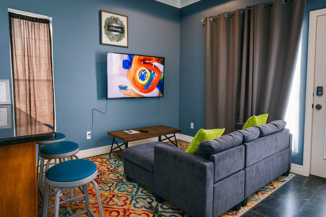 Colorful Coastal Living Room
Relax on the plush gray sofa with lime pillows facing a wall-mounted smart TV, vibrant patterned rug, and glass door to pool area. Bar stools and curtains create a cozy, beach-chic hangout.