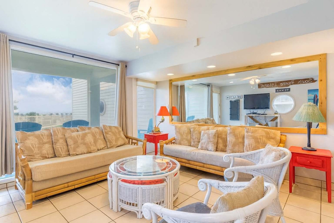 Bright Coastal Living Room with Tropical Flair & Patio Views
Relax in this airy beachside living room with bamboo and wicker furniture, tropical accents & patio views. Features include a flat-screen TV, ceiling fan, mirrored wall, and vibrant decor.
