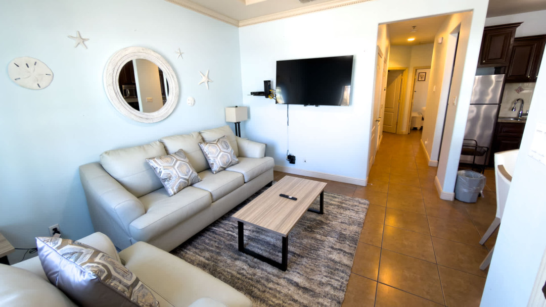 Bright, modern living space featuring cozy sofas, a sleek coffee table, and a stylish accent mirror. Enjoy an open layout flowing into the kitchen, soft lighting, and a welcoming setup ideal for relaxing, streaming, or gathering after a day out.