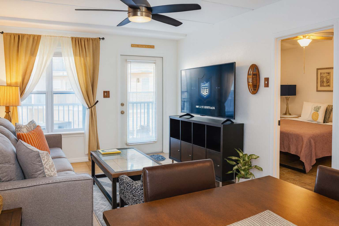 Cozy Beachfront Living Room w/ Balcony Views & Smart TV. Gray sofa, wood dining table & chairs face TV under fan, with tropical wall art, plants & sheer-draped windows to balcony. Open layout flows to bedroom—ideal for lounging, streaming & Gulf.
