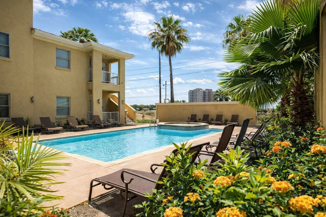 Sunlit poolside retreat framed by lush palms & vibrant blooms, offering a serene escape just steps from modern amenities. Lounge chairs line the sparkling blue water, inviting guests to unwind under open skies in a tropical, resort-style ambiance.