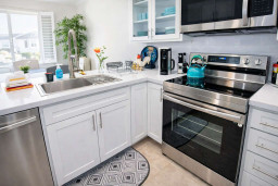 Bright, coastal-inspired kitchen featuring sleek white cabinetry, stainless steel appliances & a turquoise kettle accent. Natural light fills the space, highlighting the clean countertops, fresh florals & patterned rug—perfect for seaside charm.