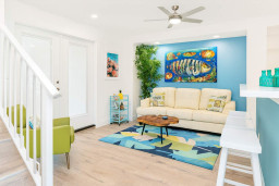 Bright coastal living room with vibrant fish art, blue accent wall, modern furnishings and colorful rug. Features open layout, natural light and beach-themed decor—perfect for relaxing and entertaining.