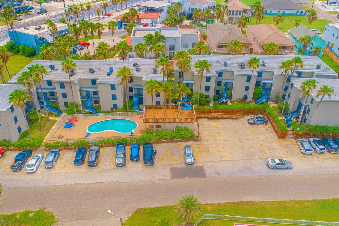 Aerial Beachfront Complex Panorama
Elevated drone view reveals a modern low-rise resort with central sparkling pool, palm-shaded parking, lush green lawns, and ocean proximity. Multi-building layout offers easy access to amenities amid swaying palms.
