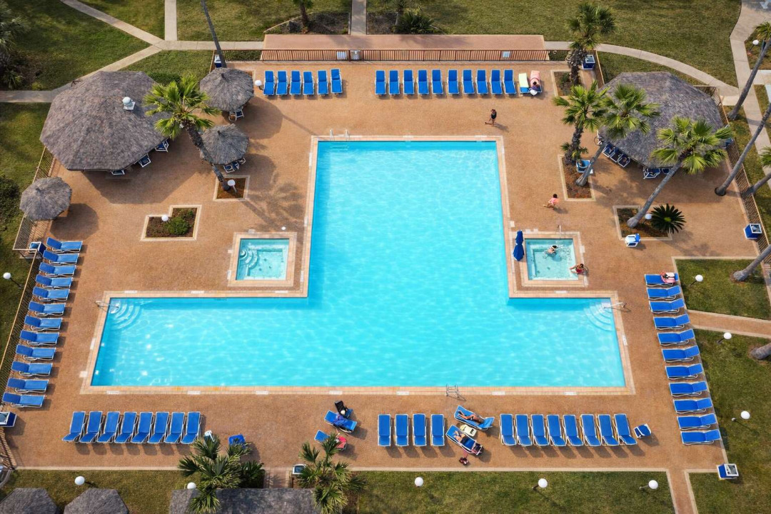 Resort-style pool retreat with connecting tubs, shuffleboard, palapa cabanas & lush tropical landscaping—perfect for relaxing under the sun or entertaining guests in a serene, well-appointed outdoor space.