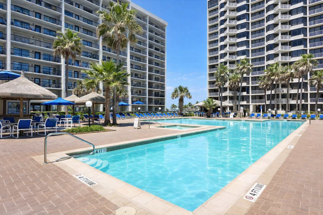Tropical resort-style pool between oceanview towers with shaded cabanas, palm trees, and lounge seating — perfect for relaxing under the sun or enjoying a sunset swim.