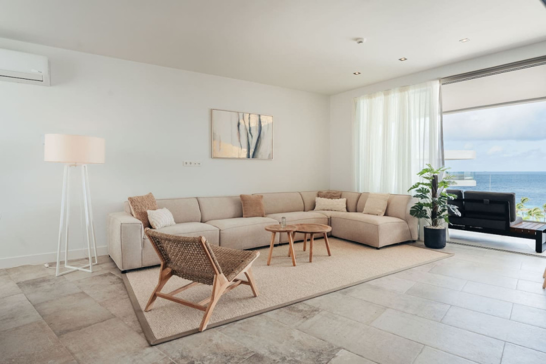 This serene living area features a cozy beige sectional, stylish woven chair, and minimalist decor, complemented by an air conditioner and expansive windows offering stunning ocean views. Perfect for relaxation.