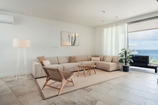This serene living area features a cozy beige sectional, stylish woven chair, and minimalist decor, complemented by an air conditioner and expansive windows offering stunning ocean views. Perfect for relaxation.