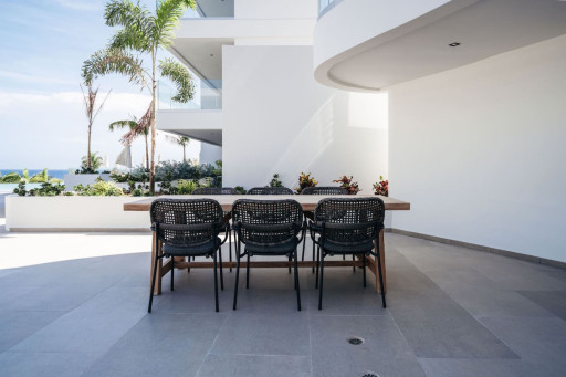 Spacious outdoor dining area with a wooden table and black woven chairs, surrounded by tropical plants and offering a clear, open sky view.