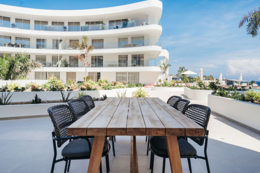 Close-up of a wooden dining table with black woven chairs, situated in an outdoor space surrounded by plants and offering a beautiful building view.